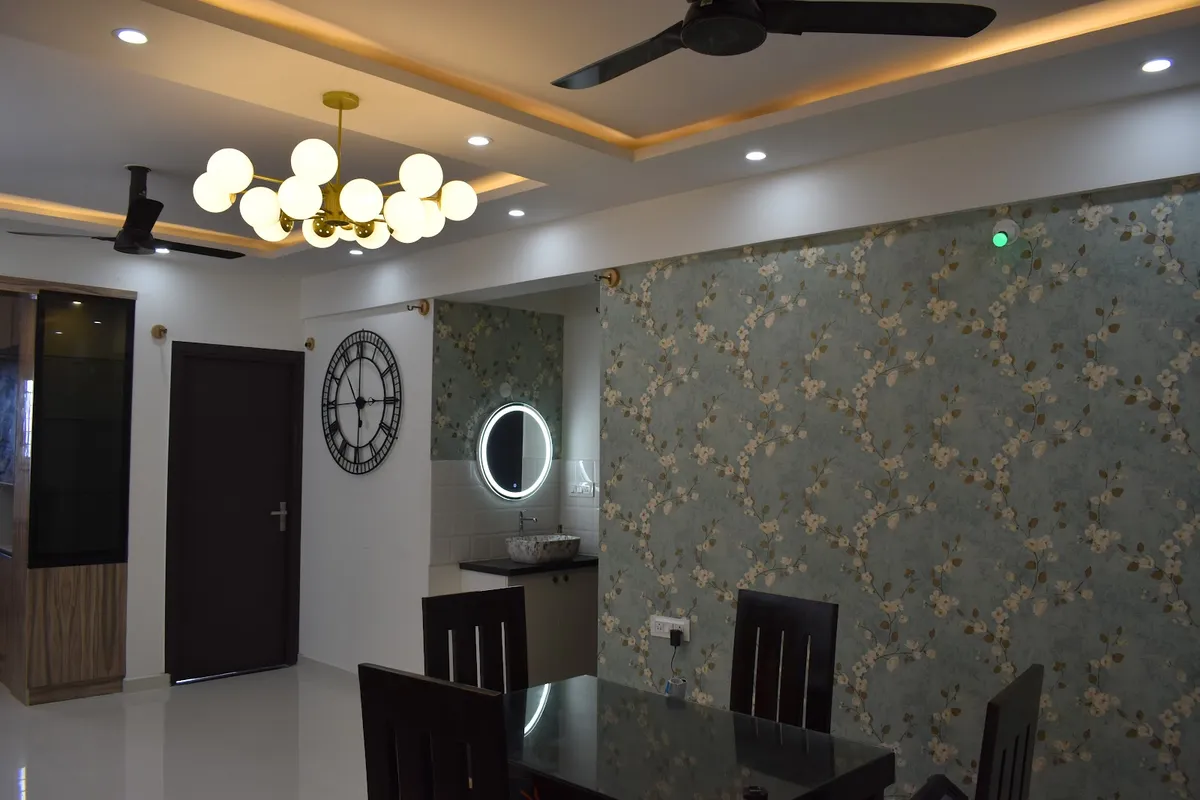 A dining room with a sage-green floral wallpaper feature wall, a black wood dining table, a brass multi-globe chandelier and a wall clock