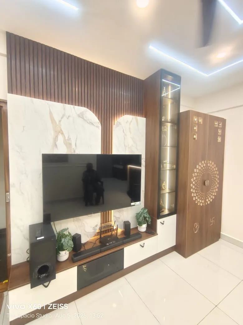 Living room TV wall with a marble slab backdrop, wood fluted panelling, a lit glass display shelf, and an adjoining wood pooja cabinet with a brass jaali motif.