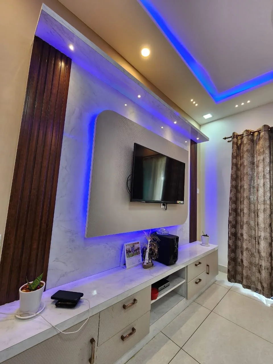 Living room TV feature wall with a marble-effect panel, wooden fluted side borders and blue cove lighting under a false ceiling.