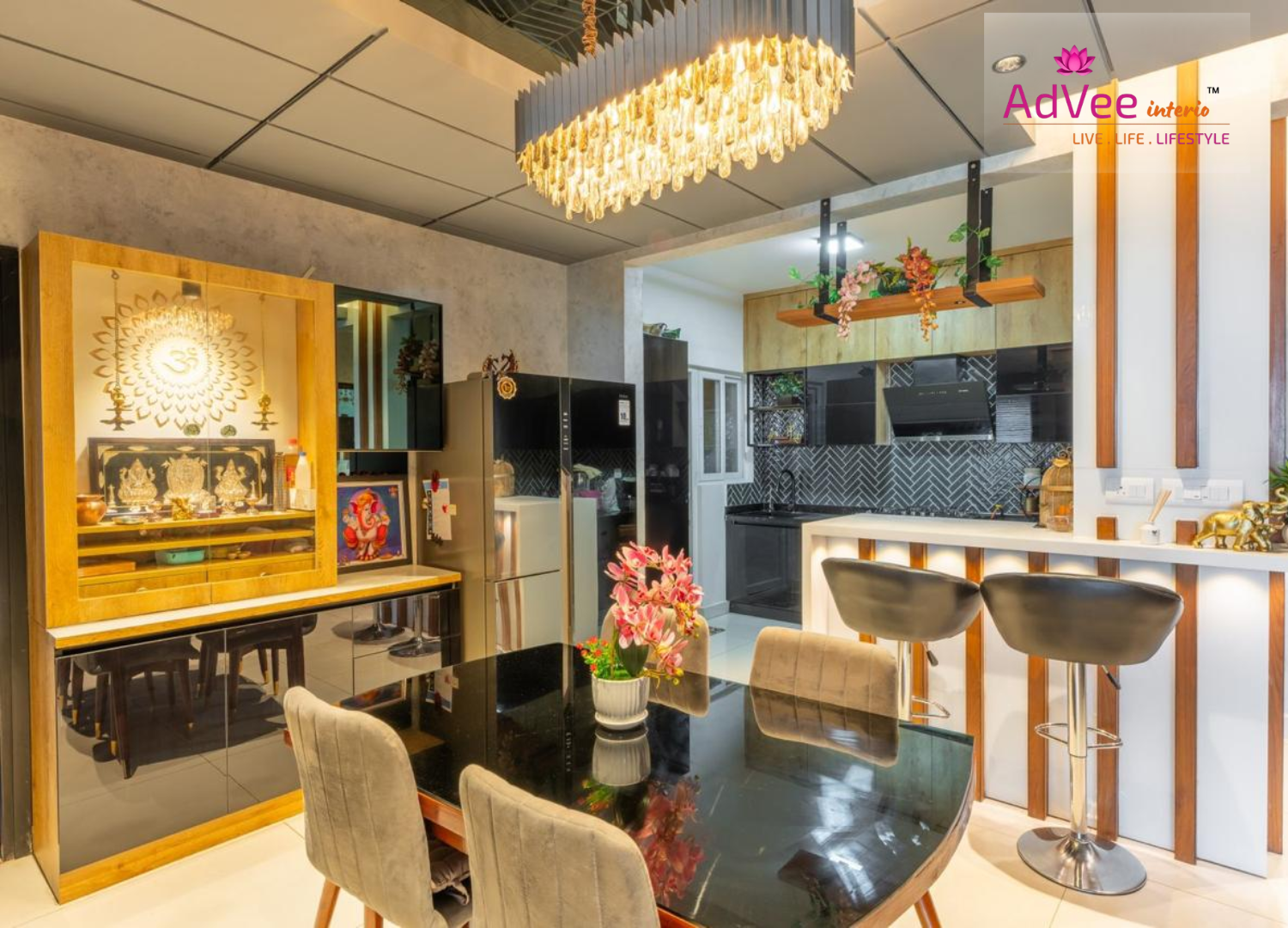 Open dining area with a black stone-top table and velvet chairs beneath an oversized crystal chandelier, a wood-framed pooja unit with a lit Om motif backdrop