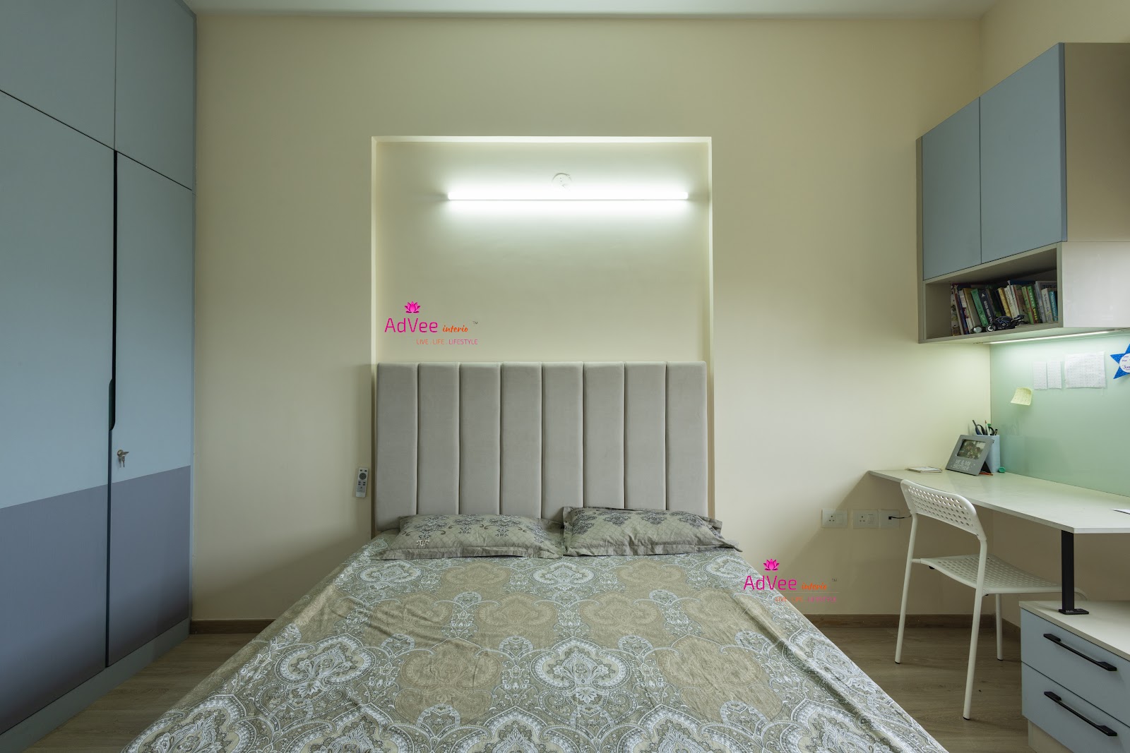 Bedroom with a channel-tufted grey headboard set into a lit wall niche, a slate-blue wardrobe on one side and a built-in study desk with overhead cabinet on