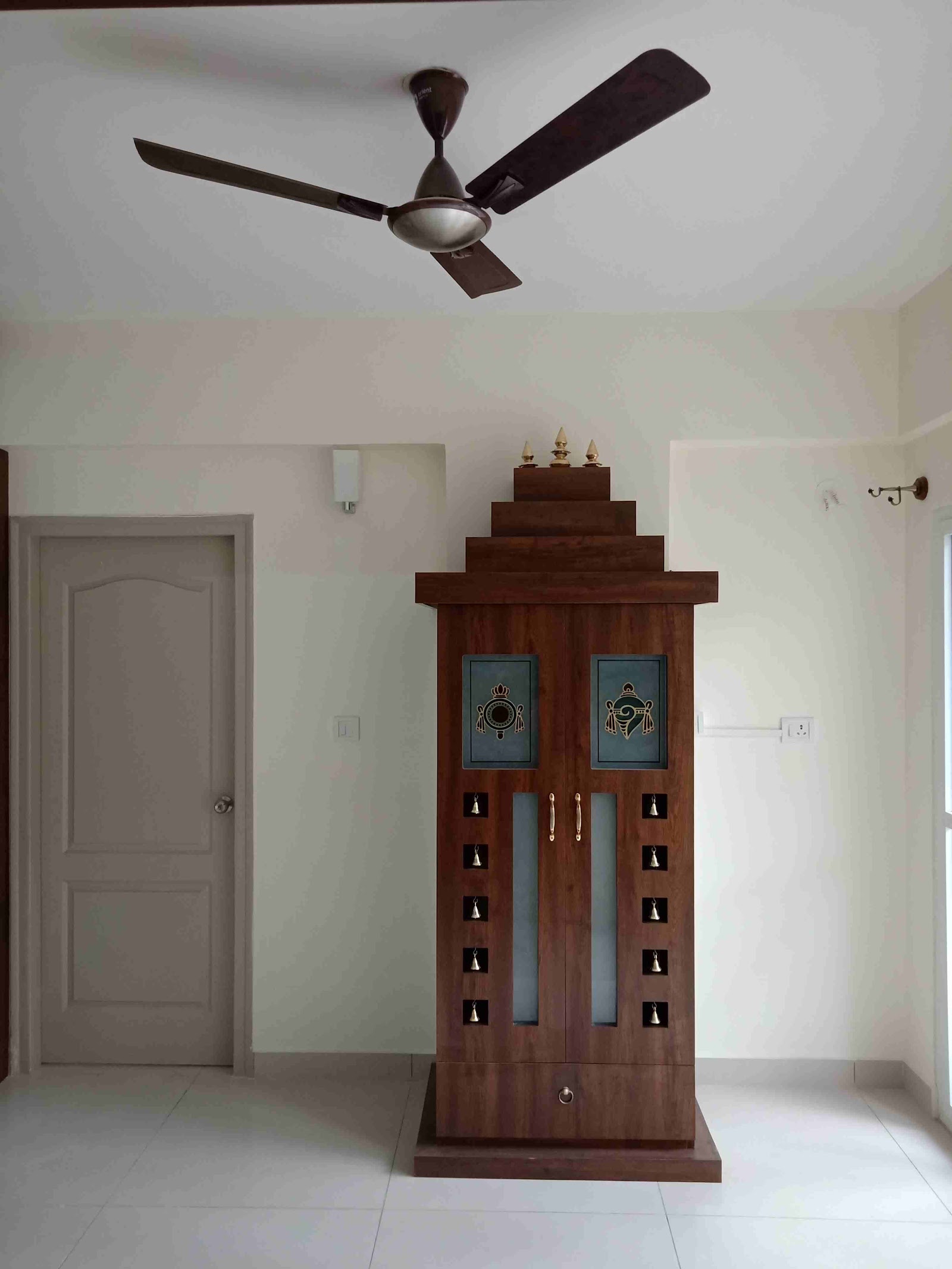 A freestanding wooden pooja unit with a tiered temple-shaped pediment, brass finials, hanging bells, and frosted glass door panels with traditional motifs.