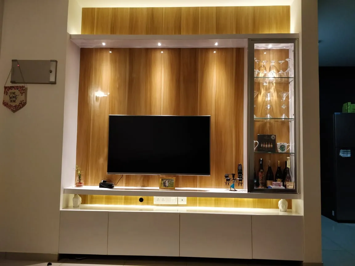 Night shot of a teak-veneer TV wall with spot-lit reveals, white gloss drawer bank below and a glass-fronted bar cabinet on the right holding upturned martini