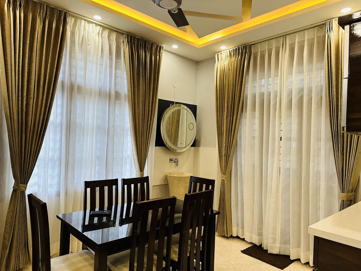 Real photo of a dining room with a dark wood table and chairs, floor-to-ceiling gold and sheer curtains, a cove-lit POP ceiling