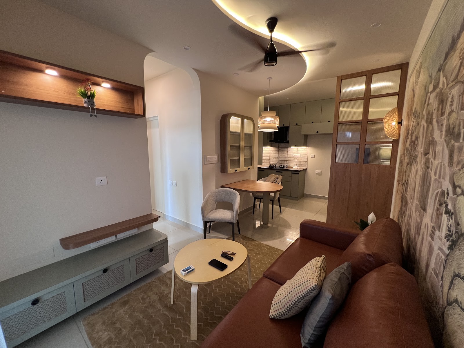 Open-plan living-dining-kitchen with a curved cove-lit ceiling, a tan leather sofa, a round dining table and sage-green kitchen cabinetry