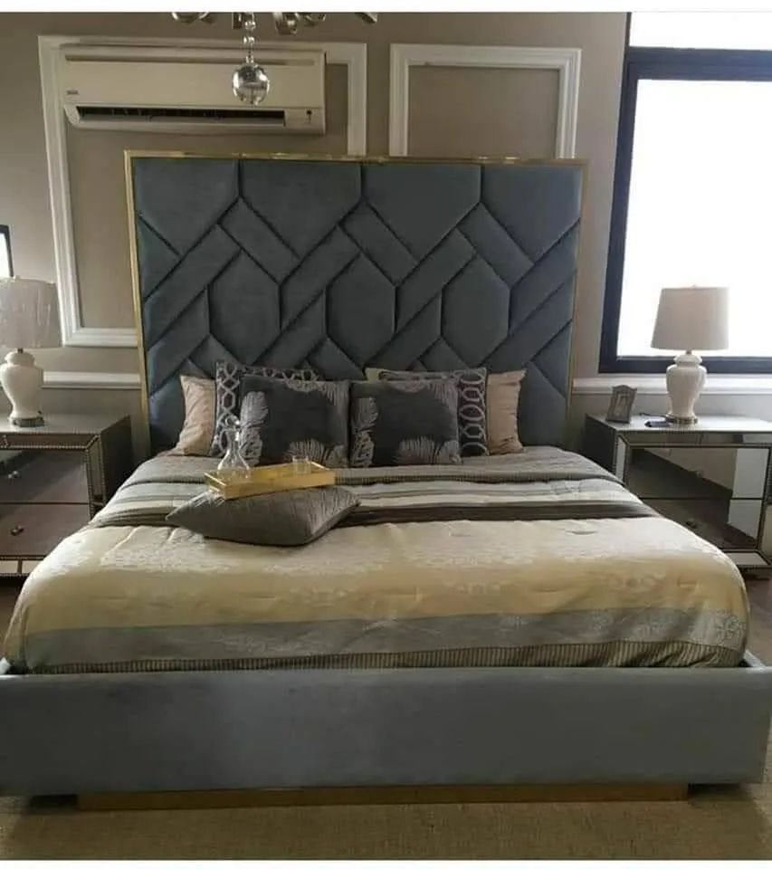 Photo of a master bedroom with a tall channel-tufted grey velvet headboard bordered in gold trim, mirrored bedside tables