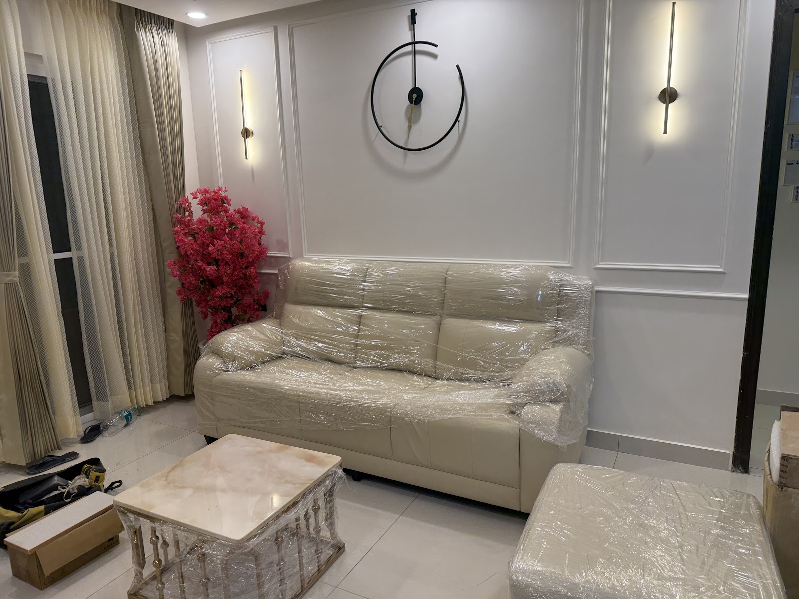 Living room with panel-moulded walls, twin linear brass wall sconces, and a beige leather sofa still wrapped for delivery beside a marble-and-brass coffee