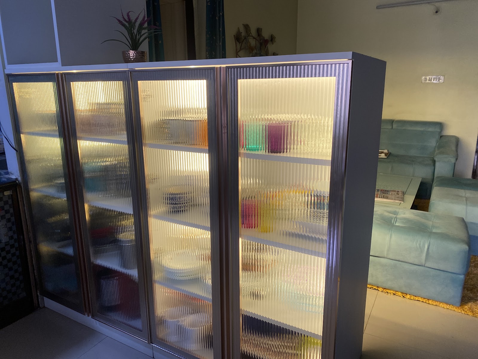 Backlit crockery display cabinet with fluted glass doors and colorful glassware, positioned in front of a mint velvet sofa set in the living room beyond.