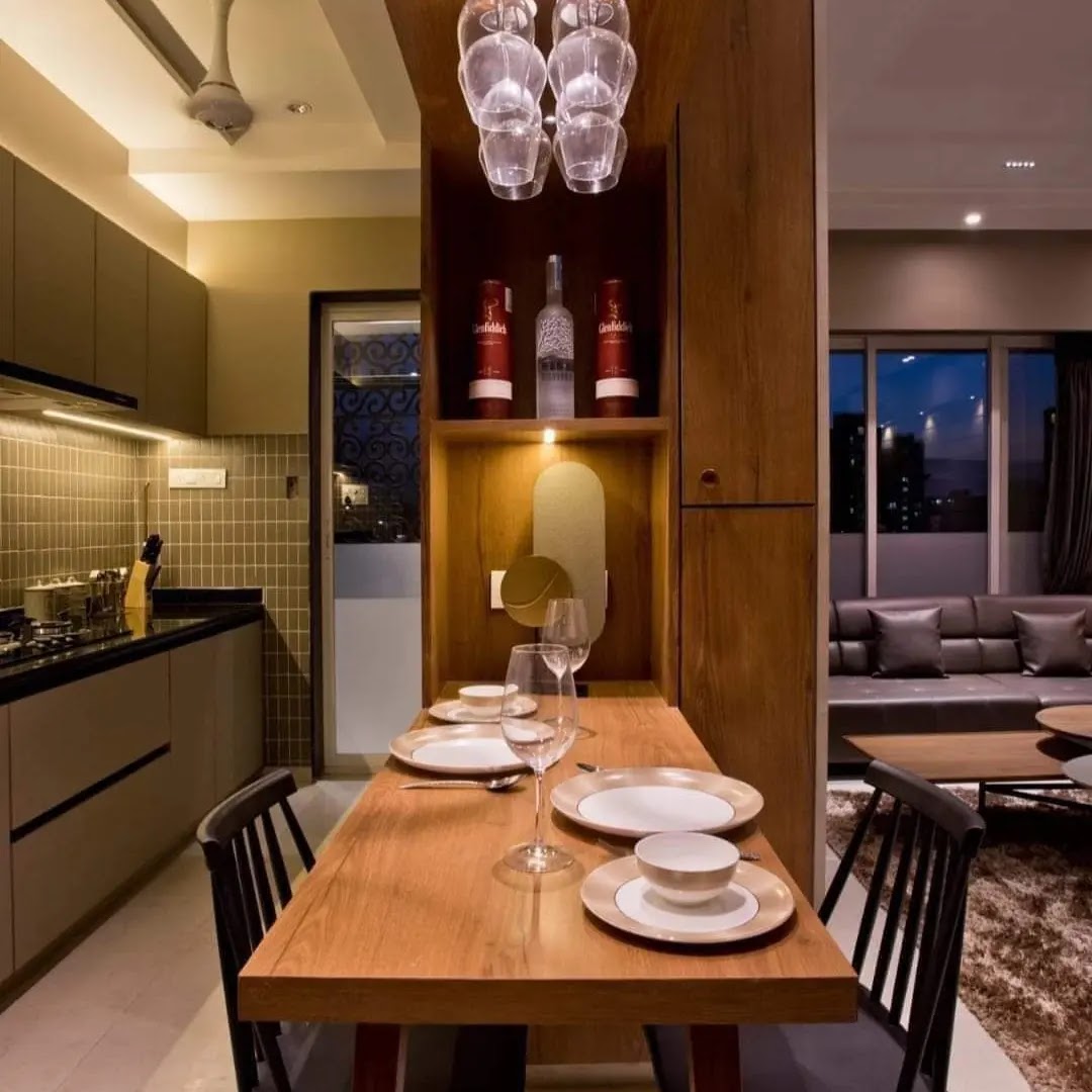 Open-plan apartment view at night showing the kitchen, a wood dining table set for two beneath a cluster glass pendant light with a built-in bar display niche