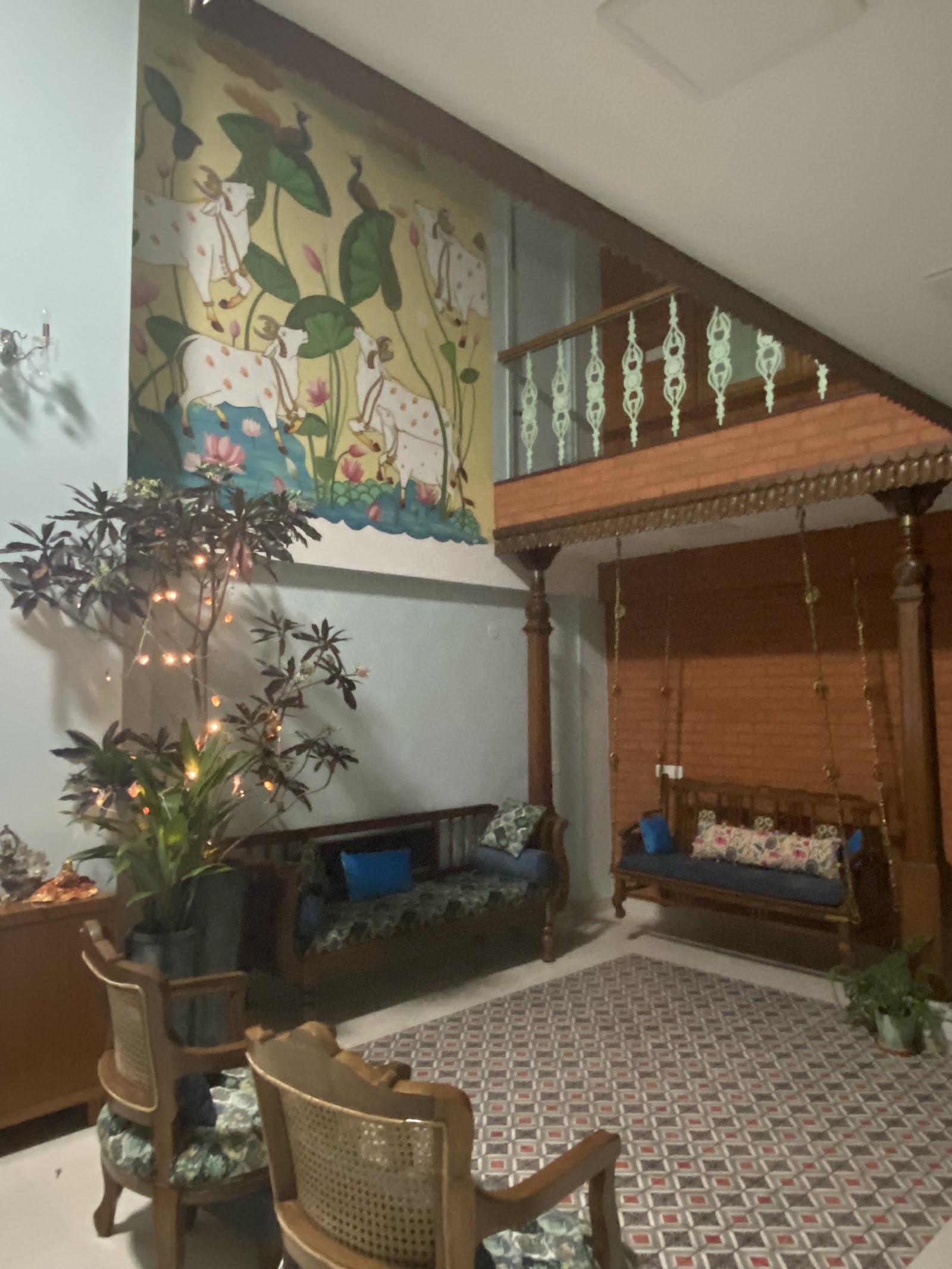 Double-height traditional sitting area with a hand-painted mural of cows and lotus flowers, a carved wooden hanging swing with blue and floral cushions