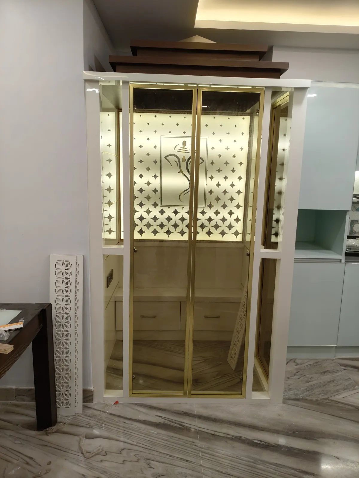 Built-in pooja unit with brass-framed etched glass doors featuring a backlit Ganesha motif, a peaked wood pediment above, and a jaali screen panel beside it.