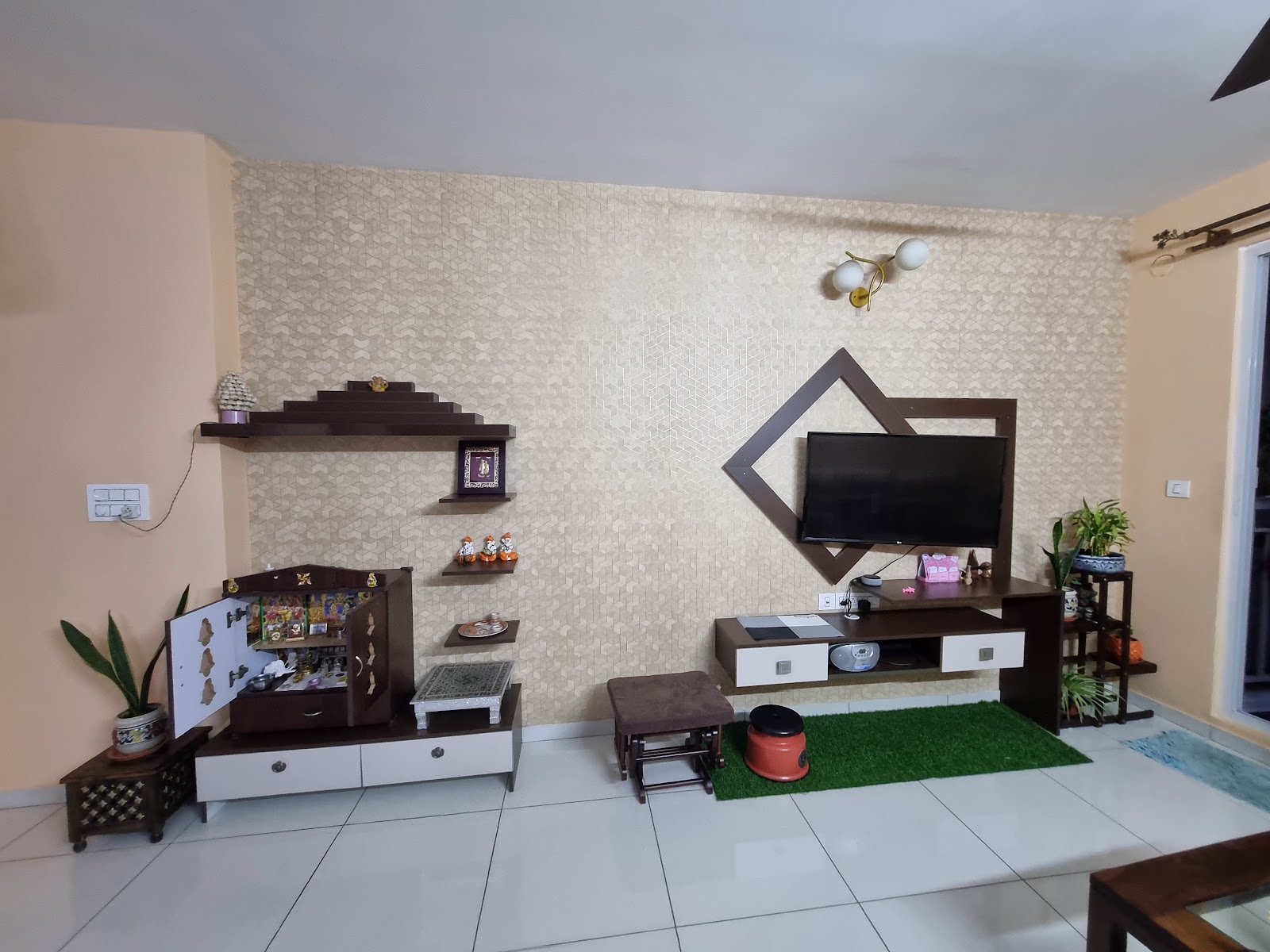 Living room with a textured geometric wallpaper feature wall, a diamond-framed wood TV console on one side and a dark-wood pooja cabinet with tiered display