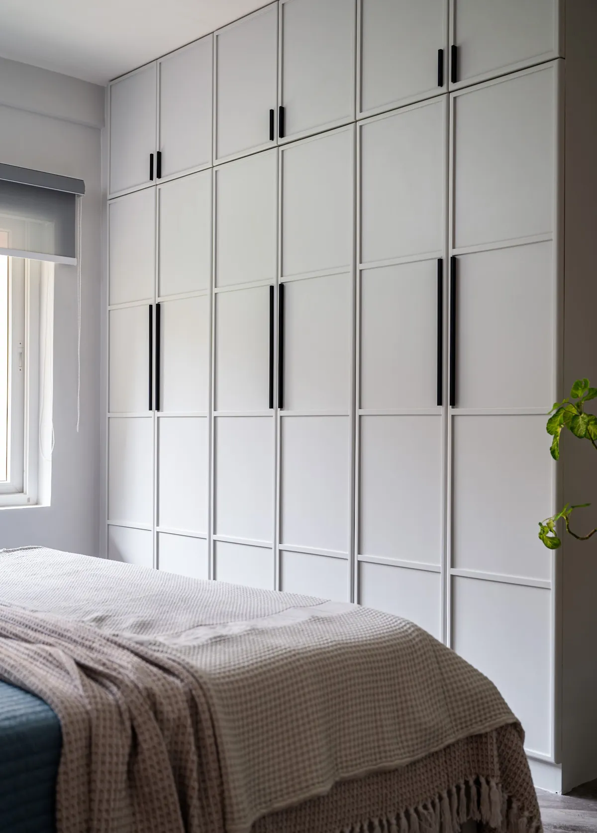 Bedroom wardrobe wall in white shaker-profile shutters running floor to ceiling with a separate loft tier and slim matt-black bar handles