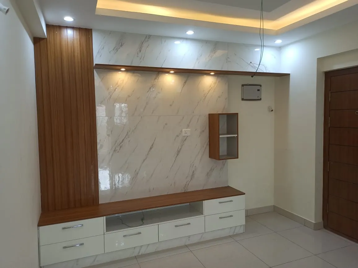 Real photo of a living room TV wall with marble-tile cladding, a fluted wood panel column, and a floating wood-and-white TV console under a cove-lit ceiling.