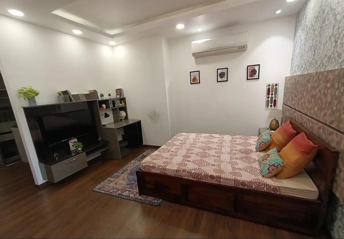 Bedroom with a wooden bed with patterned bedding and colorful cushions, a floral wallpaper headboard wall, and a dark TV unit against wood flooring