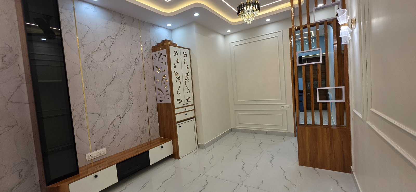 Real photo of an empty living room with a book-matched marble TV feature wall inlaid with brass strips, a compact wall-hung pooja unit with jaali-motif doors