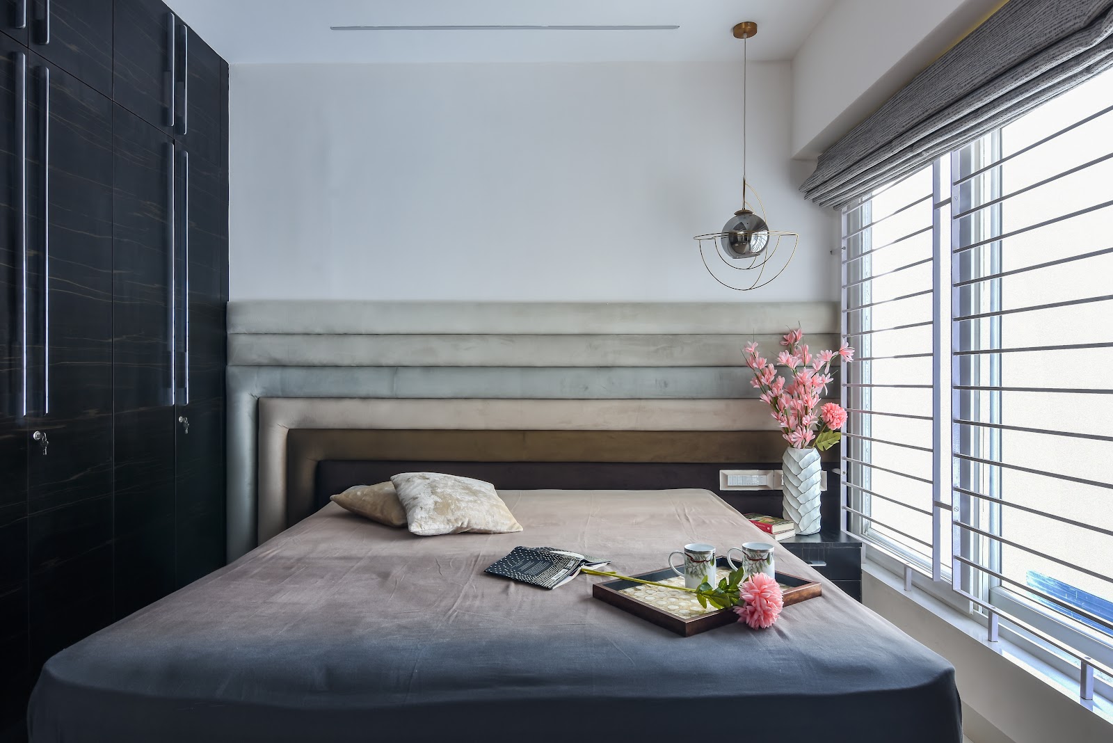 Bedroom with a channel-tufted grey velvet headboard, dark high-gloss wardrobe, and a sculptural orbital pendant light beside the window.