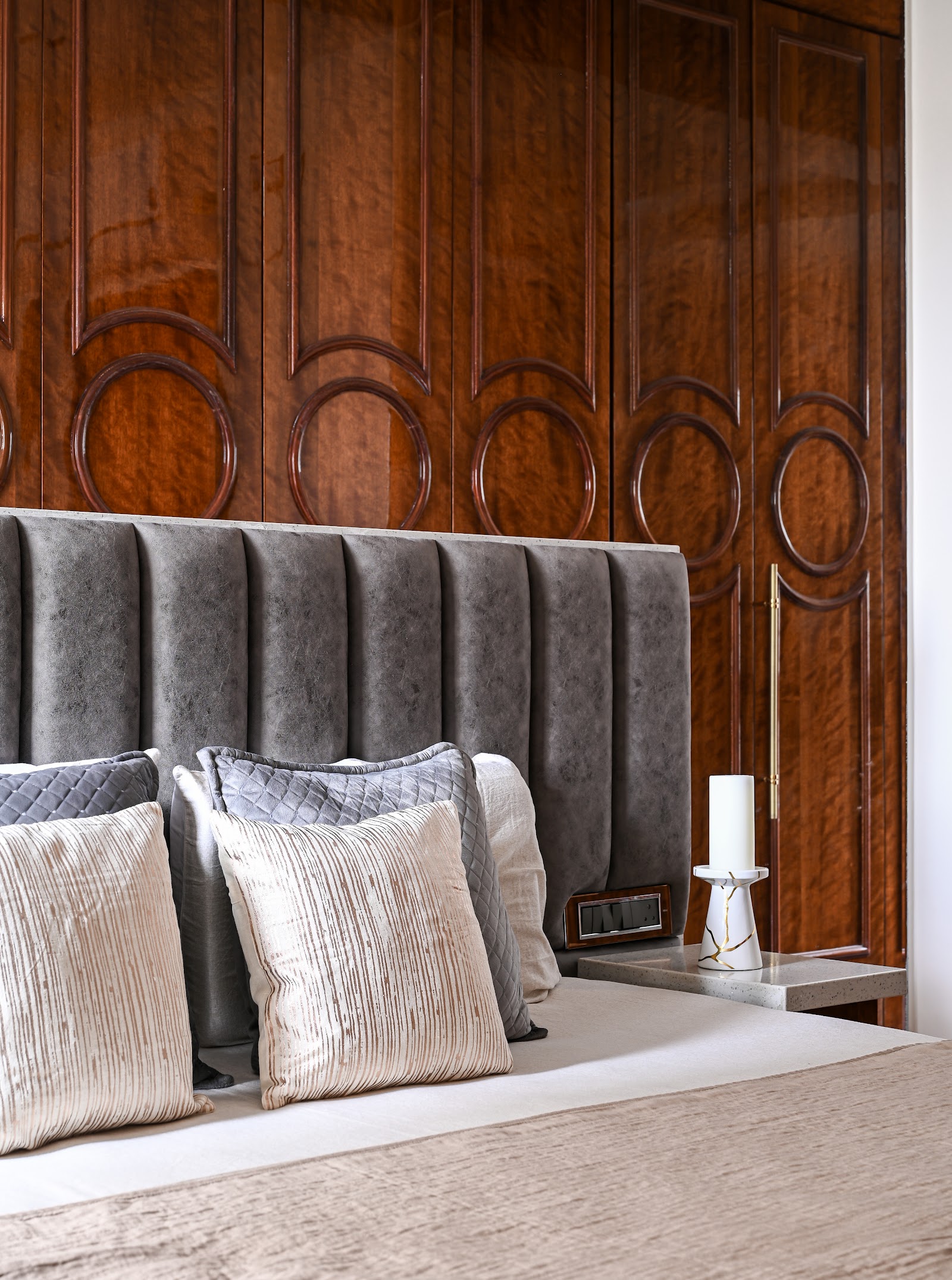 Bedroom close-up of a channel-tufted grey headboard with layered cushions against a dark wood wardrobe featuring carved circular panel mouldings