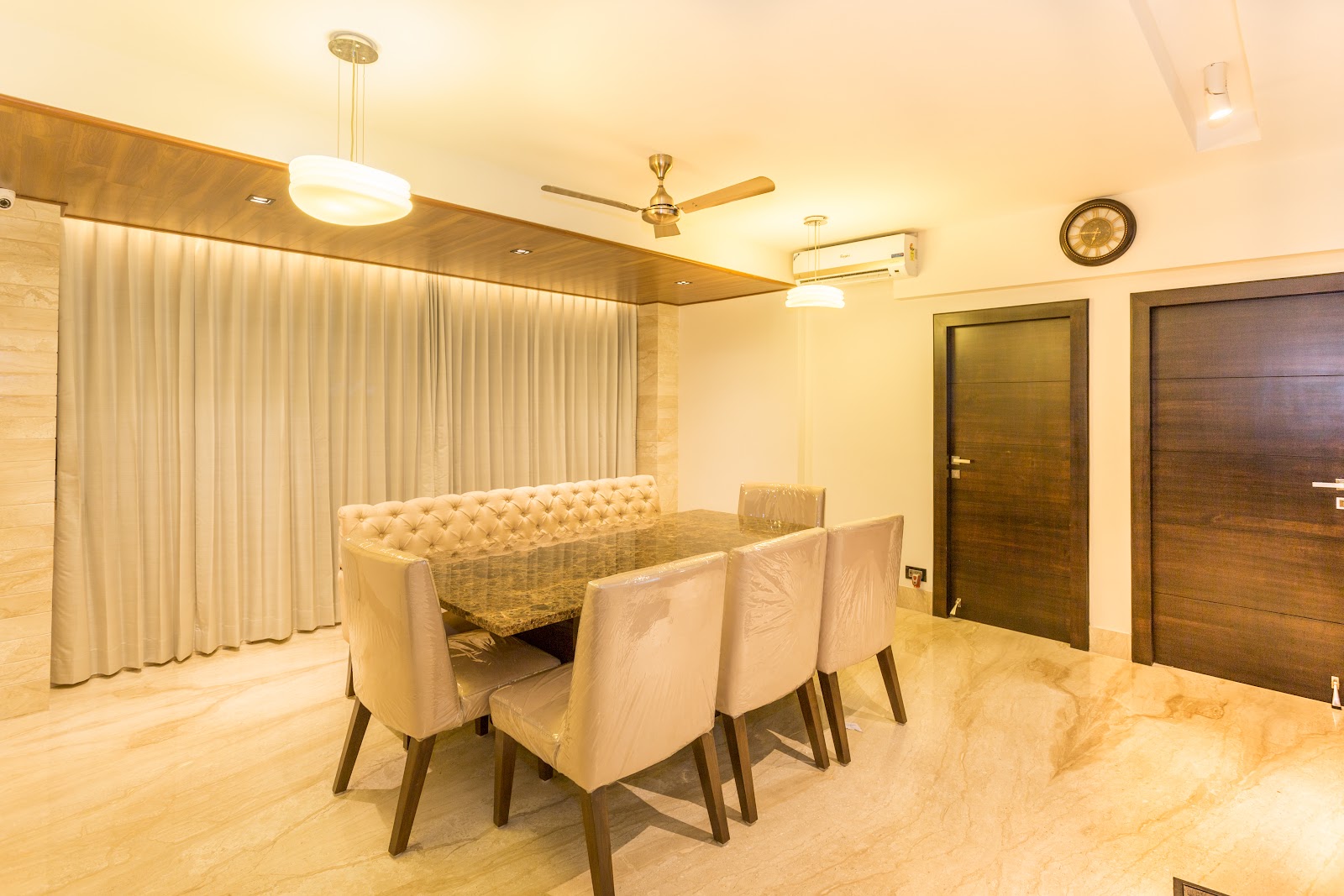 A dining room with a marble-top table and tufted bench seating under a wood-clad drop ceiling with pendant lights, chairs still under delivery plastic wrap.