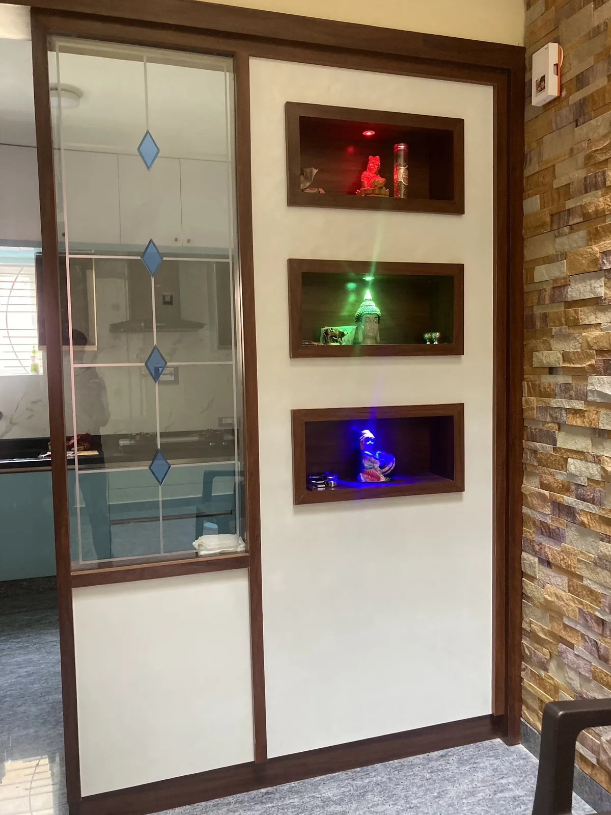 Real photo of a pooja wall with three dark-wood-framed backlit display niches holding deity idols