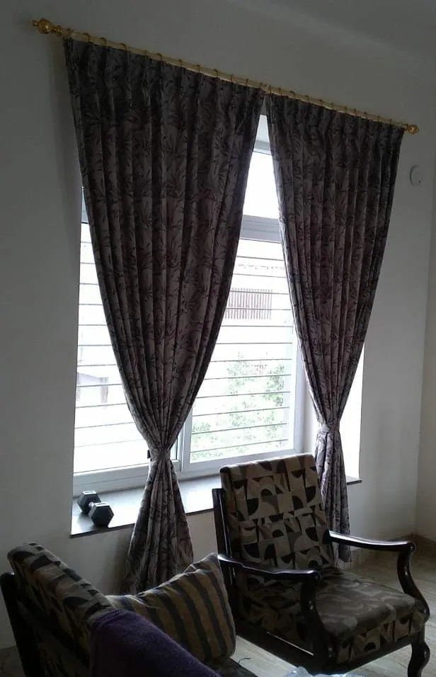 Living room corner with floral-patterned floor-length curtains on a brass pole, a carved wood armchair and sofa beside a grilled window.