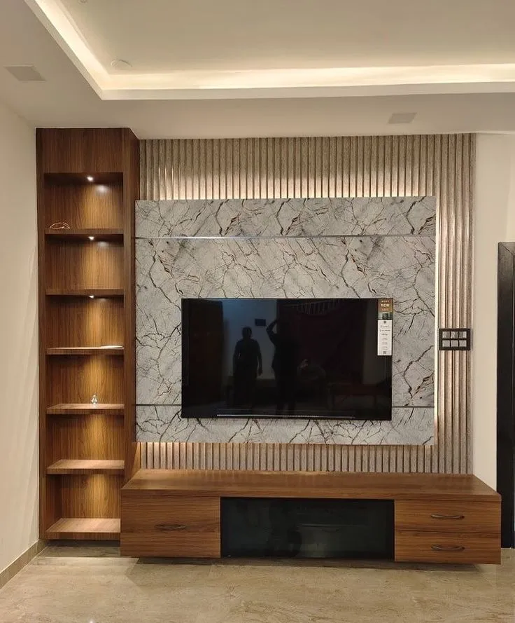 Living room TV wall with a book-matched marble slab backdrop framed by fluted brass-toned panelling, beside a lit wood open-shelf display unit.