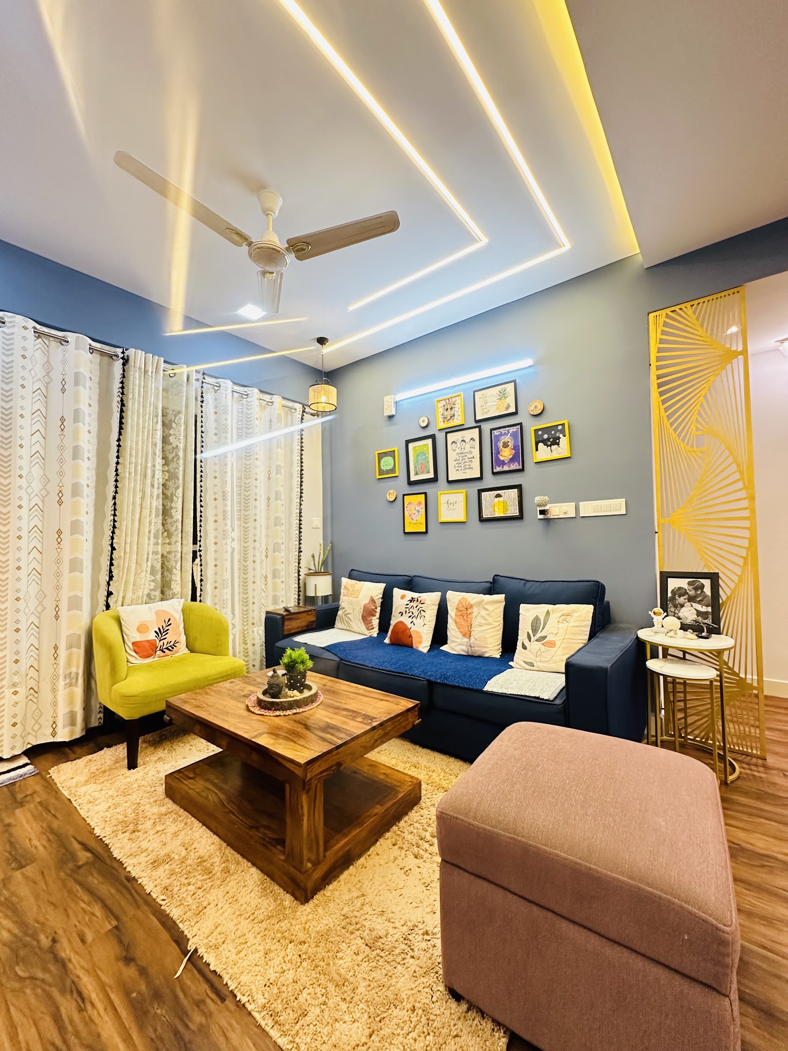 Living room with a navy sofa, mustard accent chair and a curated gallery wall of colorful framed art beneath cove-lit ceiling channels