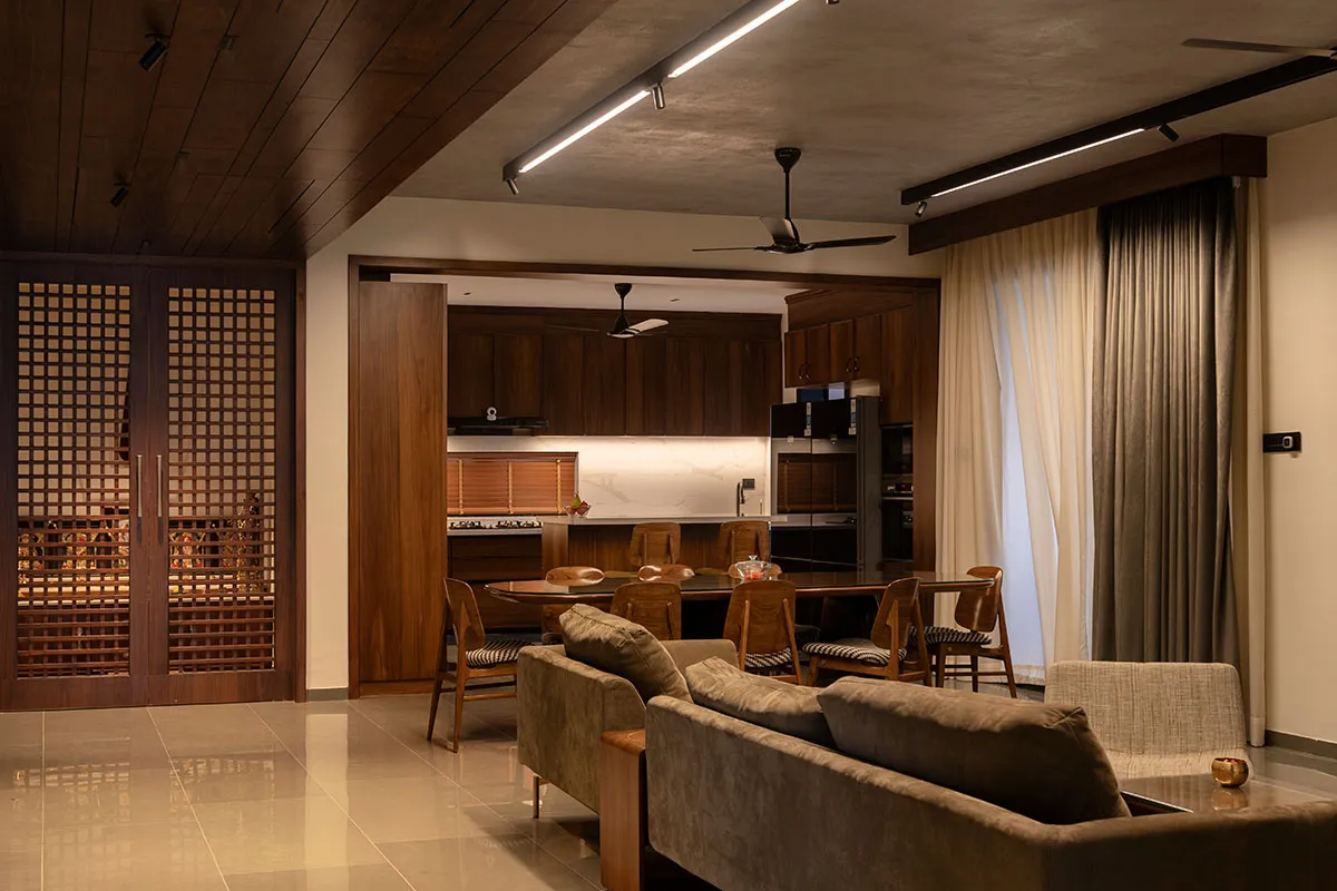 Open-plan living-dining-kitchen with a dark-wood jaali lattice door on the left, a wood-panelled ceiling, dining chairs around an oval table