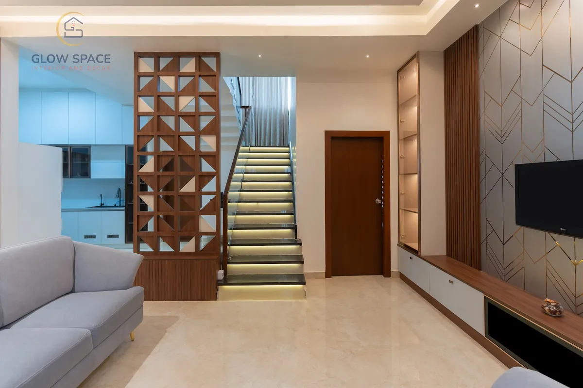 Duplex living room where a teak triangular-lattice jaali screens the white kitchen and a stone staircase with LED-lit risers climbs beside a TV wall of