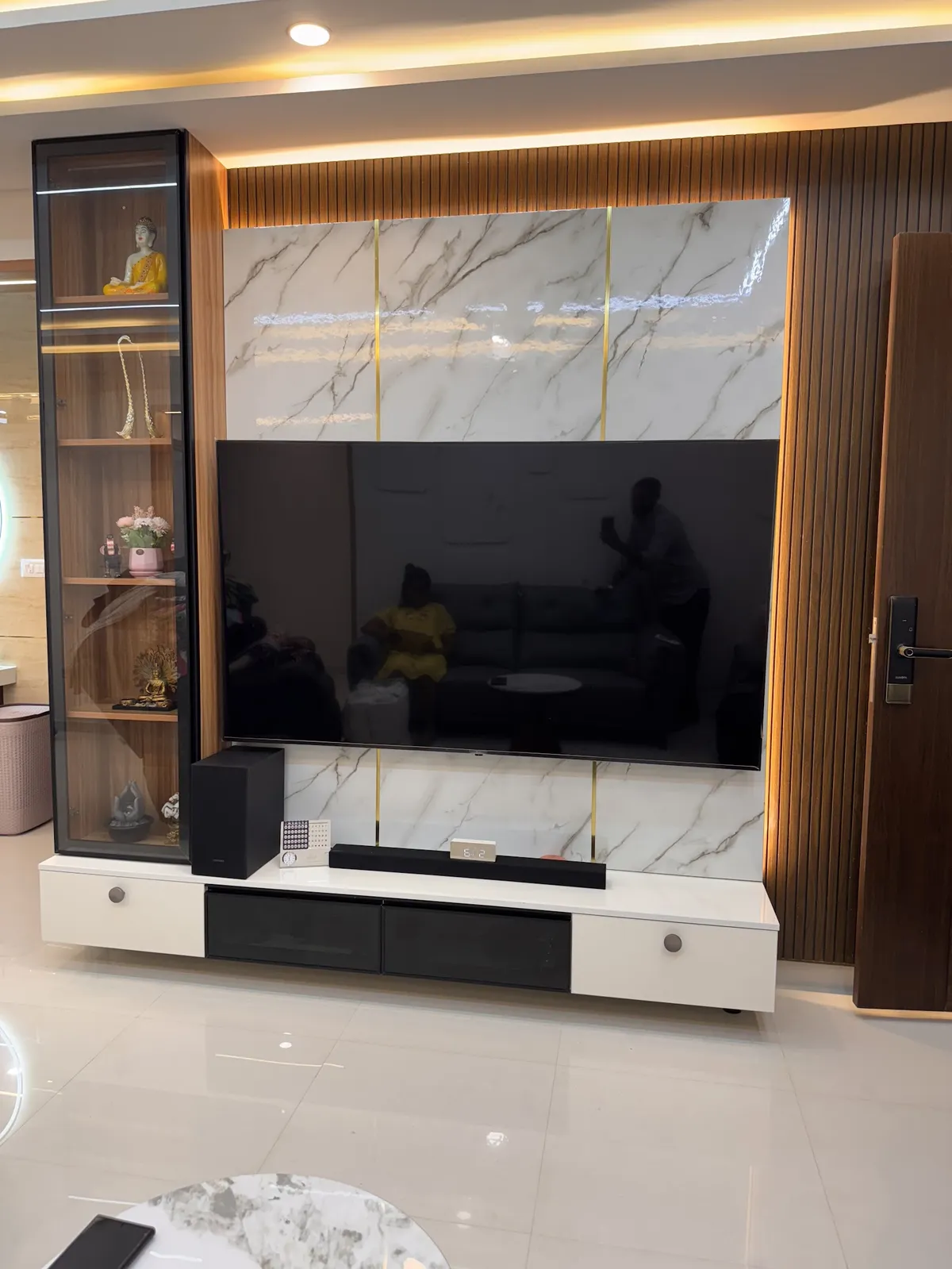 Living room TV wall in white marble-look slabs split by thin brass inlay strips, framed by fluted teak panelling and a slim black-framed glass display column