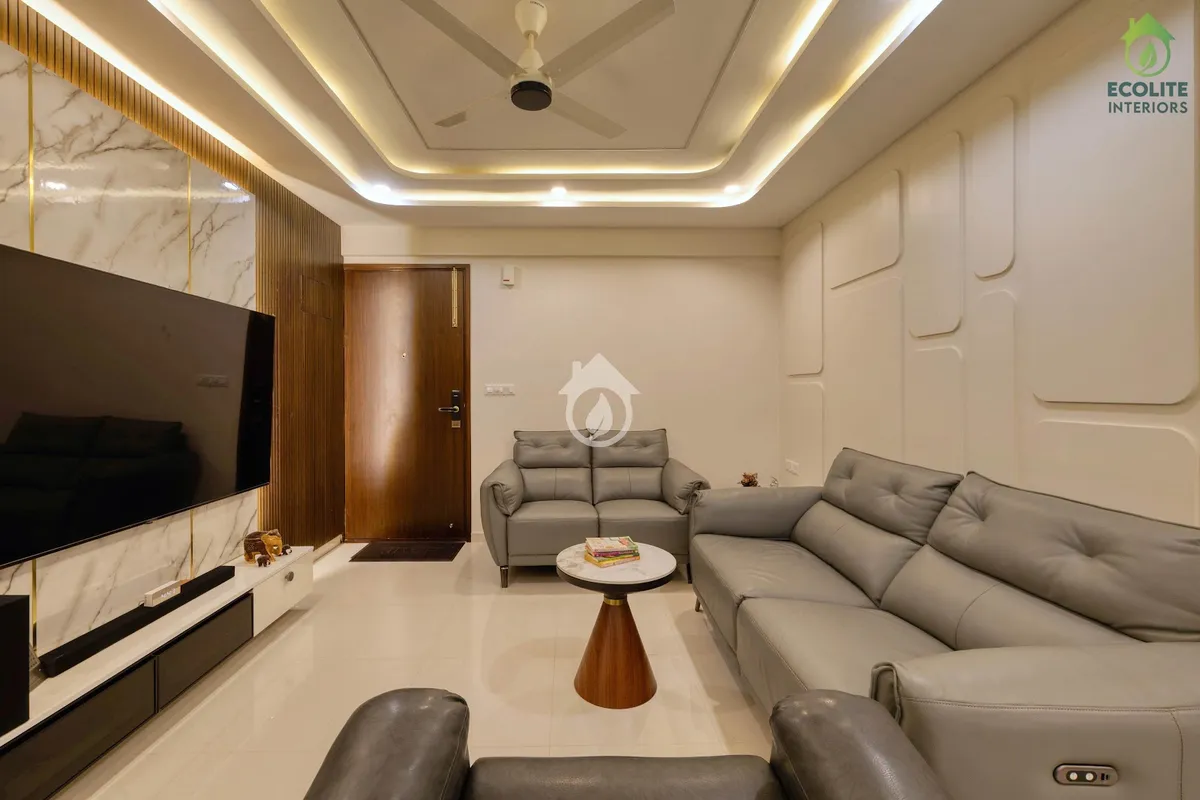 Living room with grey leather recliner sofas facing a TV wall of white marble split by brass inlays and fluted teak, a white moulded panel wall opposite