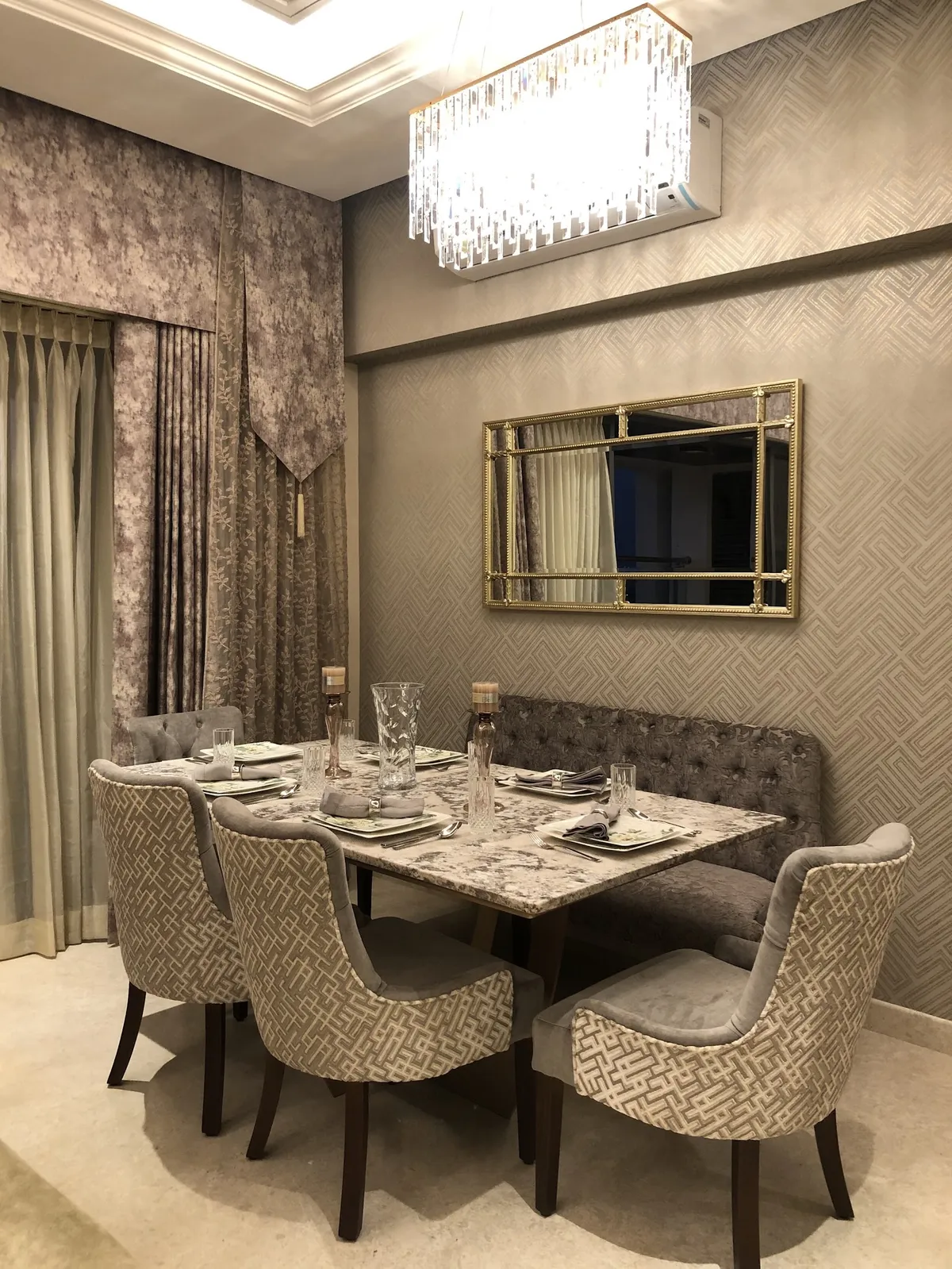 Dining nook with a marble-top table, tufted bench seating against geometric wallpaper, a gold ornate mirror, and a crystal box chandelier.