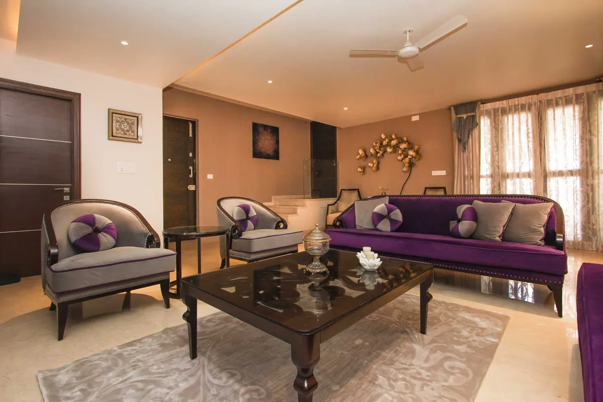 Formal living room with a purple velvet nailhead sofa facing two grey tub chairs, a heavy dark-wood glass-top table on a grey damask rug