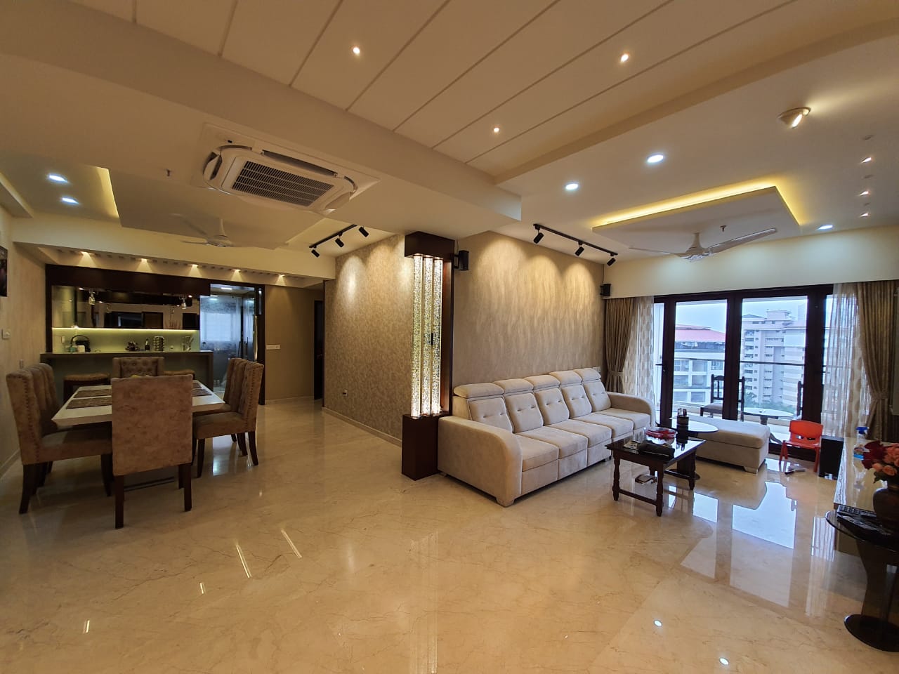 Open-plan living and dining space with cream marble flooring, a cove-lit stepped ceiling, a beige sofa set by floor-to-ceiling windows