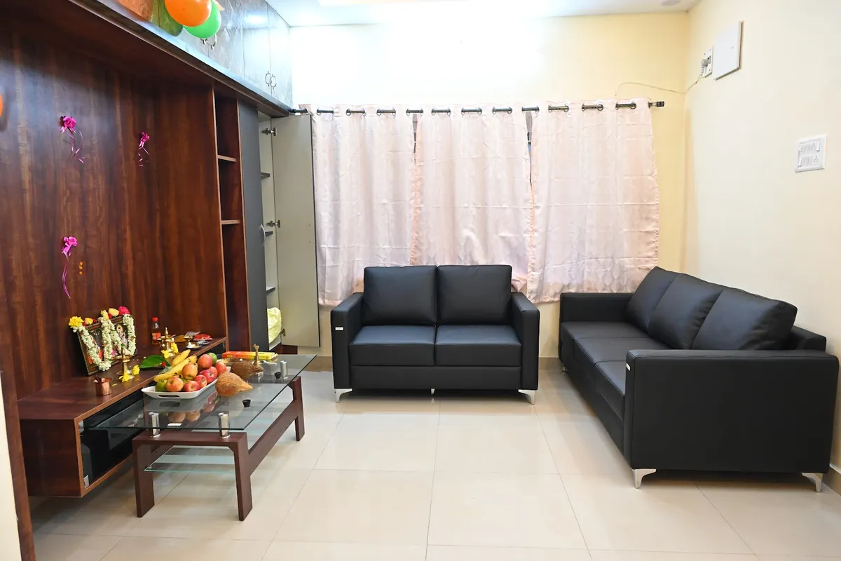 Plainly furnished living room set up for a housewarming puja, with garlanded deity photos and fruit offerings laid out on the dark wood wall unit and glass