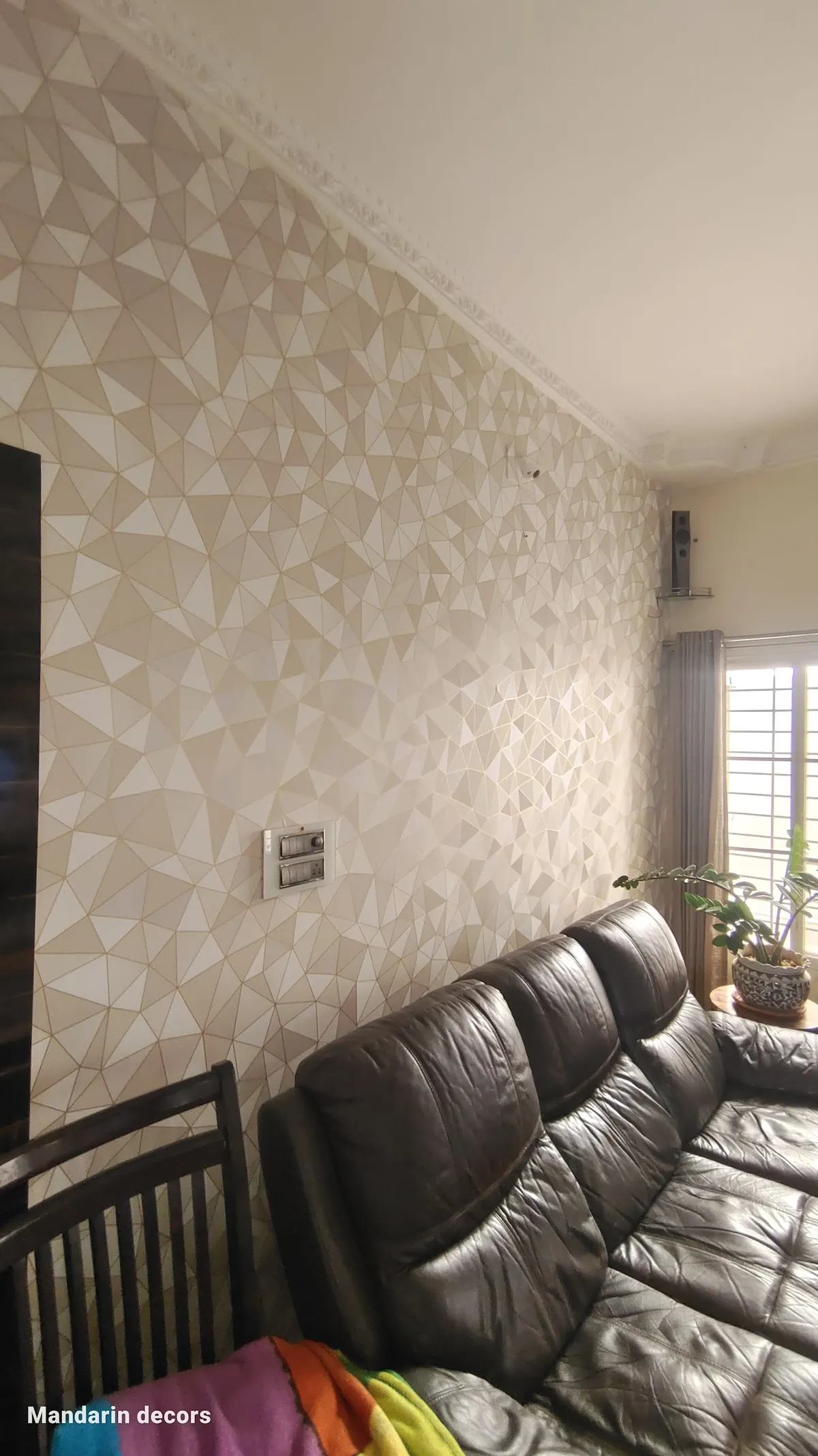 A living room with a faceted 3D geometric wallpaper feature wall, a dark brown leather sectional sofa, and sheer-and-blackout curtains by a grilled window.
