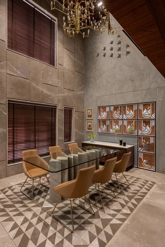 Real double-height dining room with marble-clad walls, a glass-top table on a sculptural stone base, tan leather chairs with gold legs, a brass chandelier