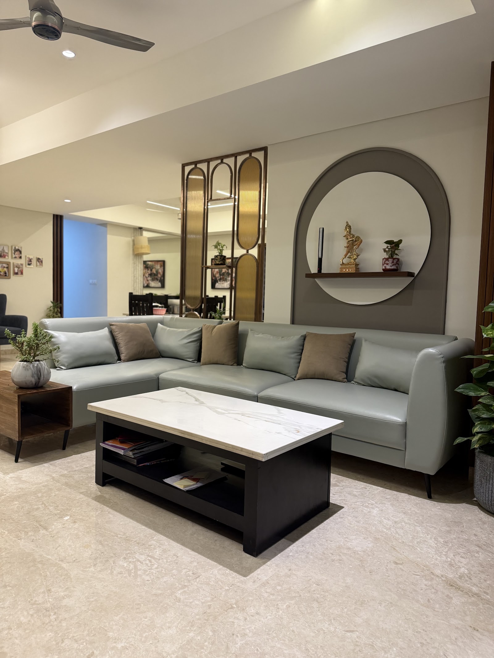 Living room with a grey leather sectional sofa and marble-top coffee table, a brass-framed reeded-glass screen beside a circular wall niche displaying a brass