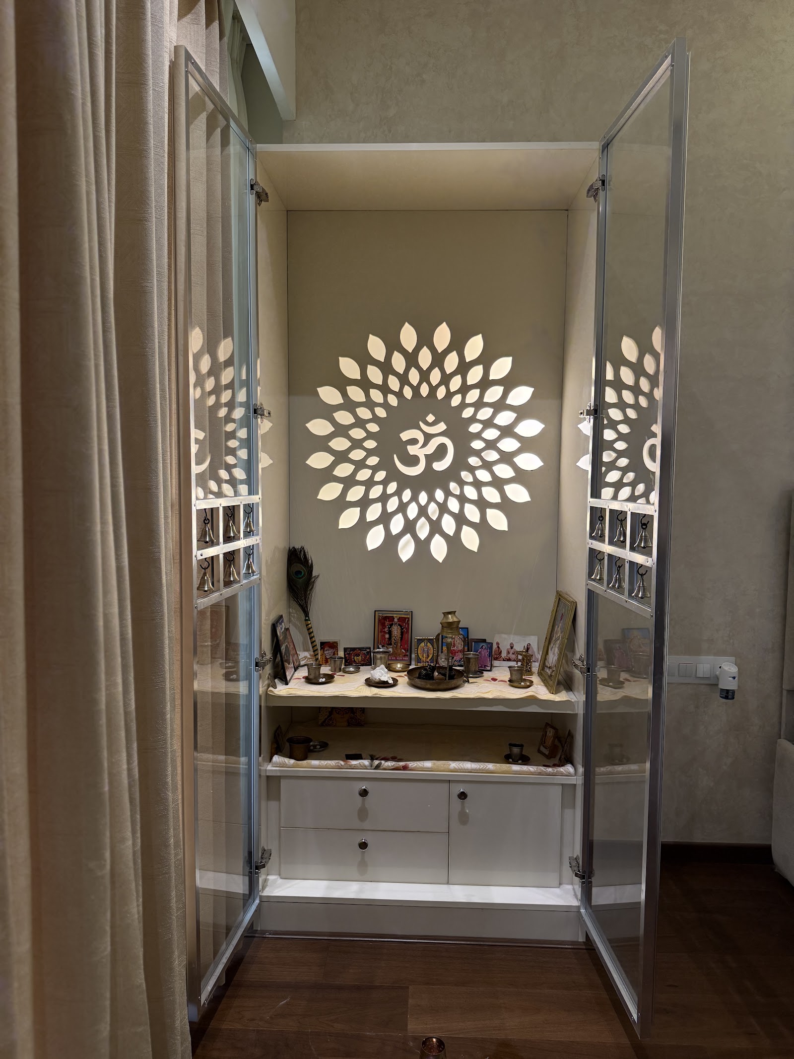 Built-in pooja unit with glass doors, brass temple bells on the door frames, a backlit lotus-and-Om wall motif