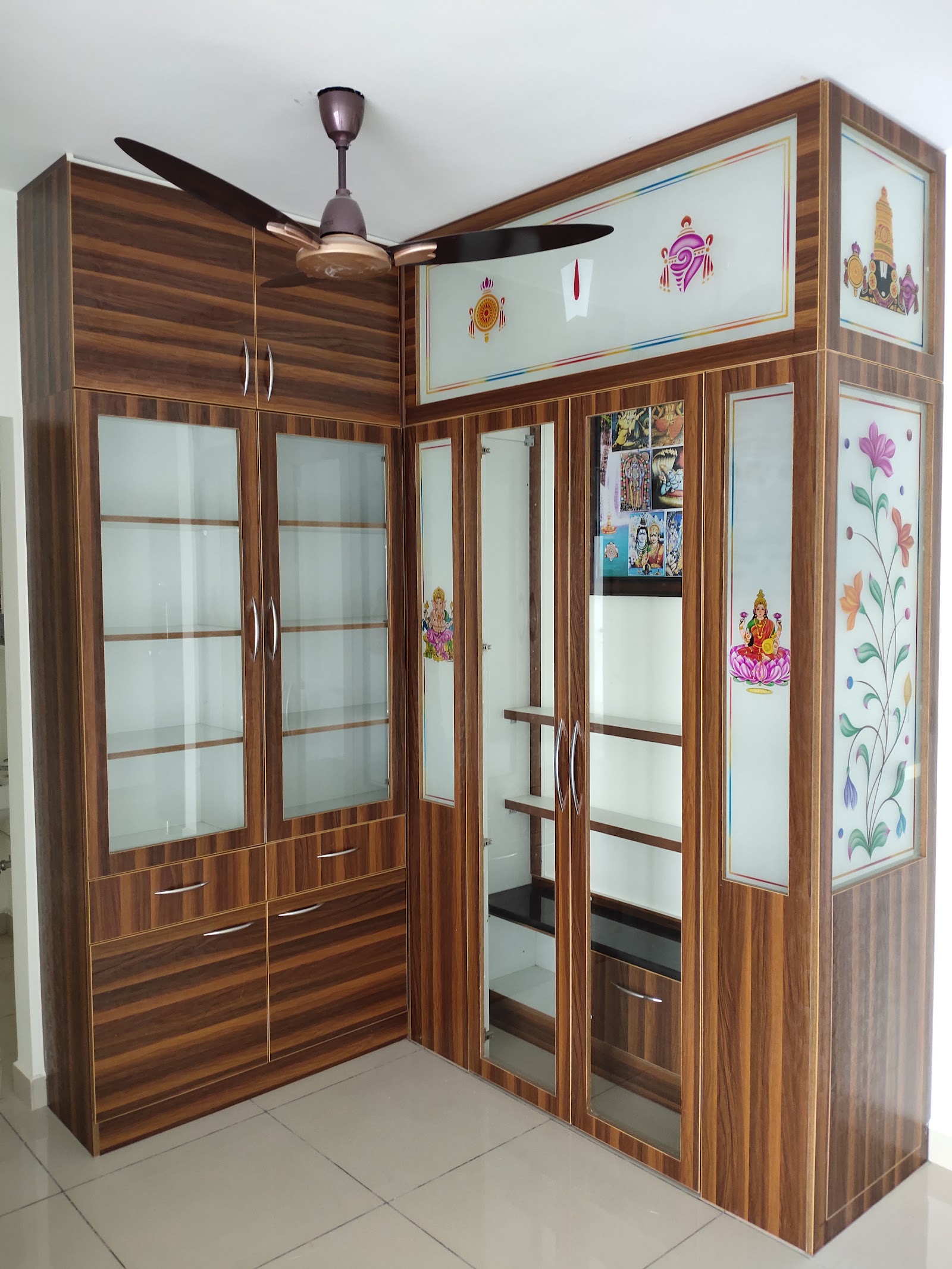 Corner wooden pooja unit with printed deity motifs on frosted glass shutter panels and open display shelving beside it.