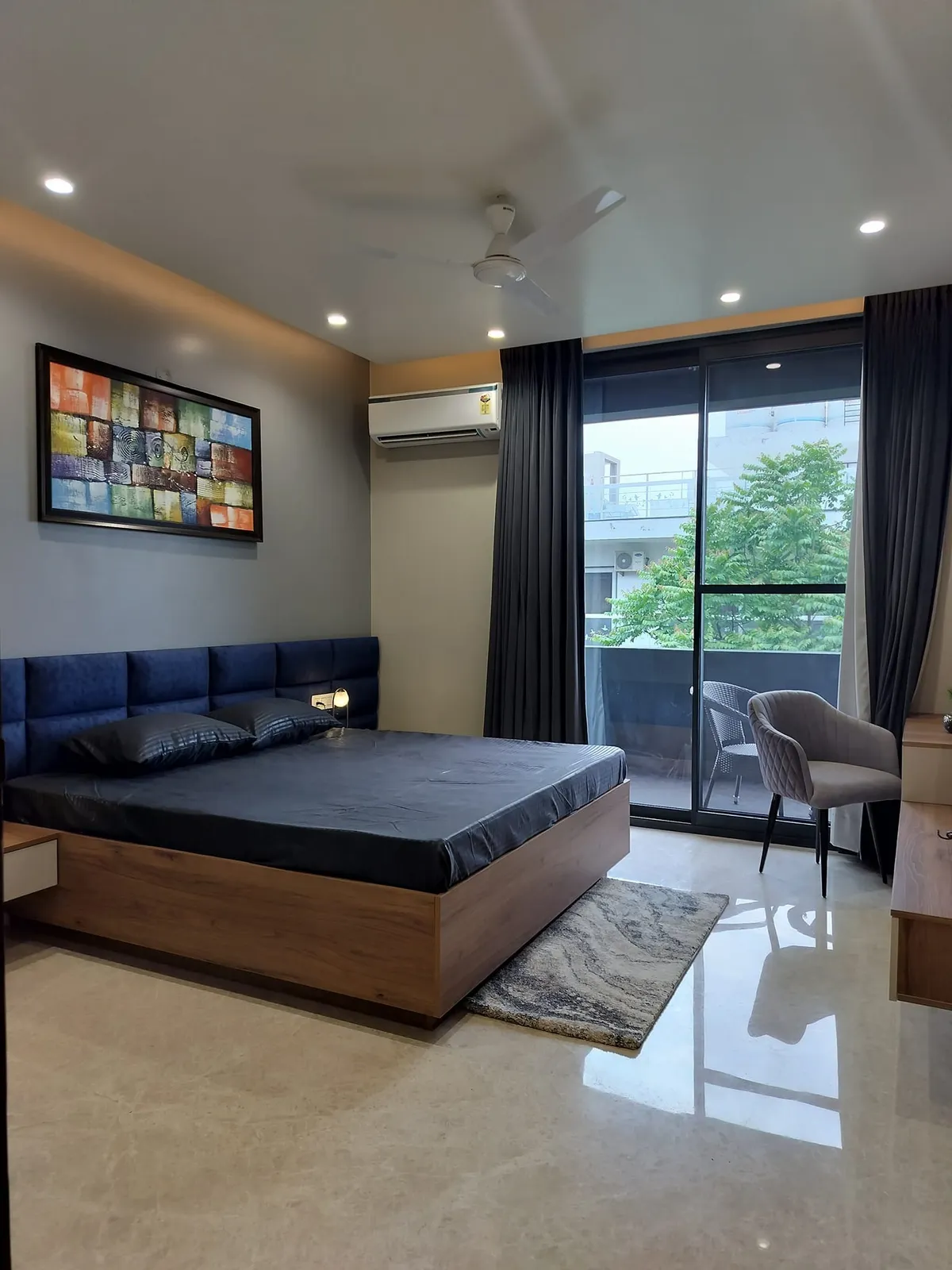 Bedroom with a navy tufted headboard on a wood platform bed, a colourful abstract wall art piece, and a glass door opening to a balcony.