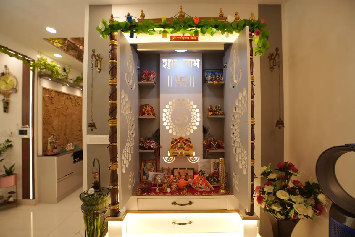 Dedicated pooja unit with carved white jaali cabinet doors, gold-trimmed pillars, a floral garland, and idols arranged on illuminated shelves.