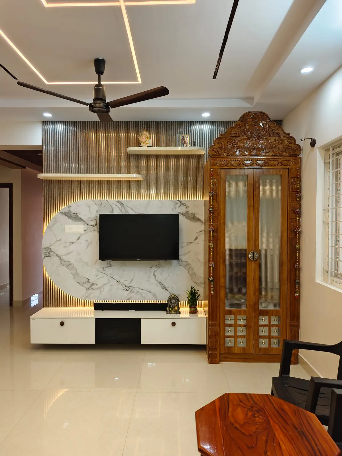 Living room with a marble and fluted-metal TV feature wall beside a hand-carved wooden pooja cabinet with glass doors and brass bells