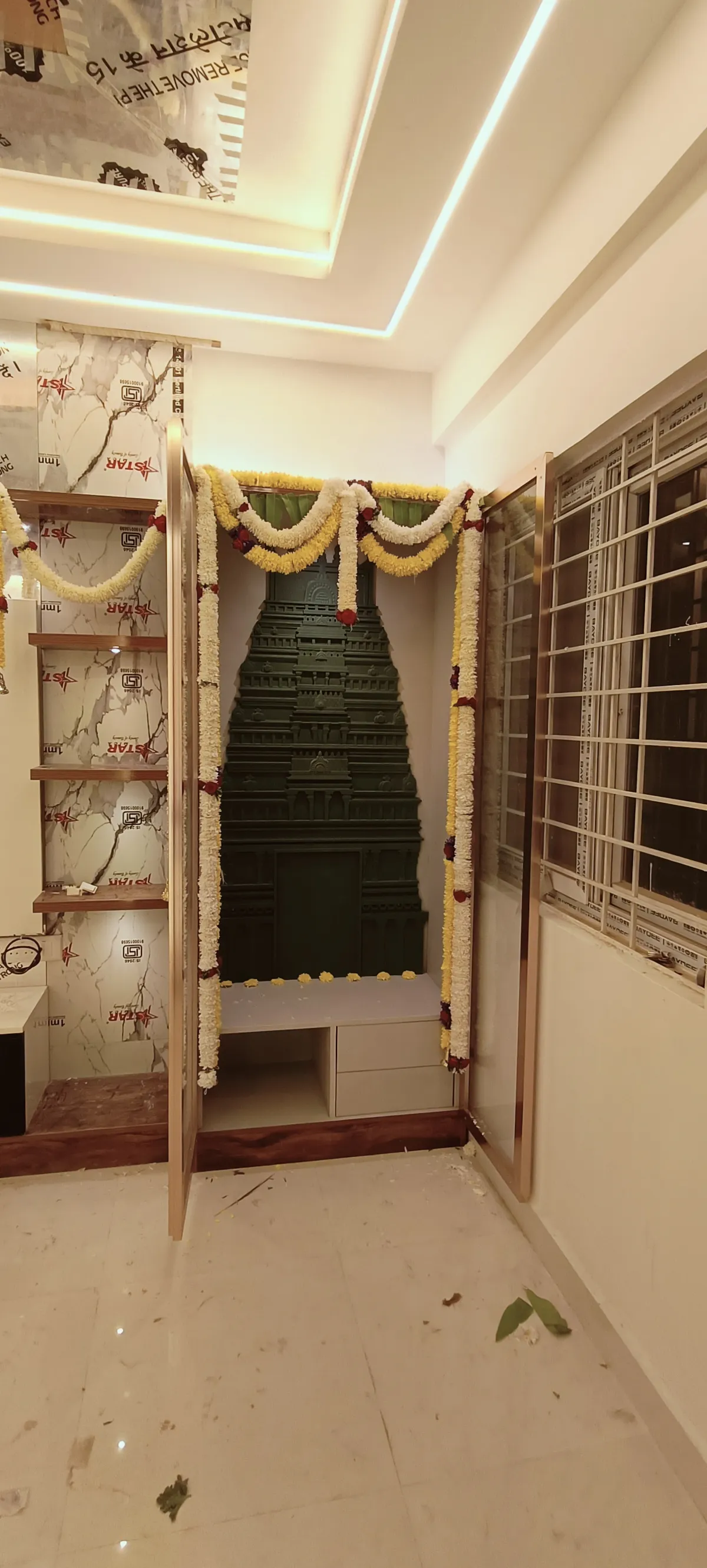 Dark-green carved gopuram mandir seated in a rose-gold framed niche, marigold garlands still draped over the opening and protective film not yet peeled off the
