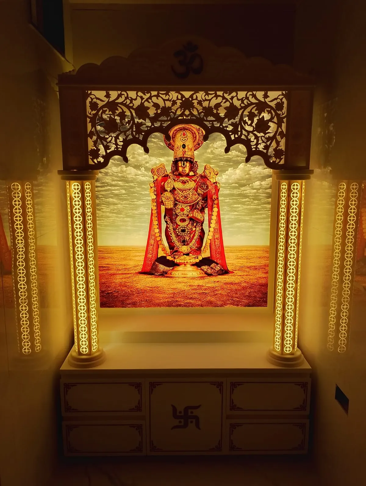 Home mandir with turned columns lit from within through CNC-cut acrylic strips, a scalloped floral jaali valance and an Om crest above a printed Balaji