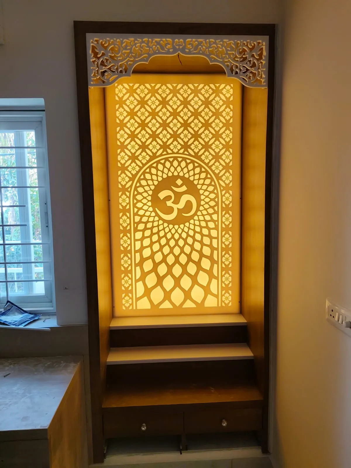 Floor-to-ceiling built-in mandir in a window corner, its backlit CNC jaali panel carrying an Om inside an arch