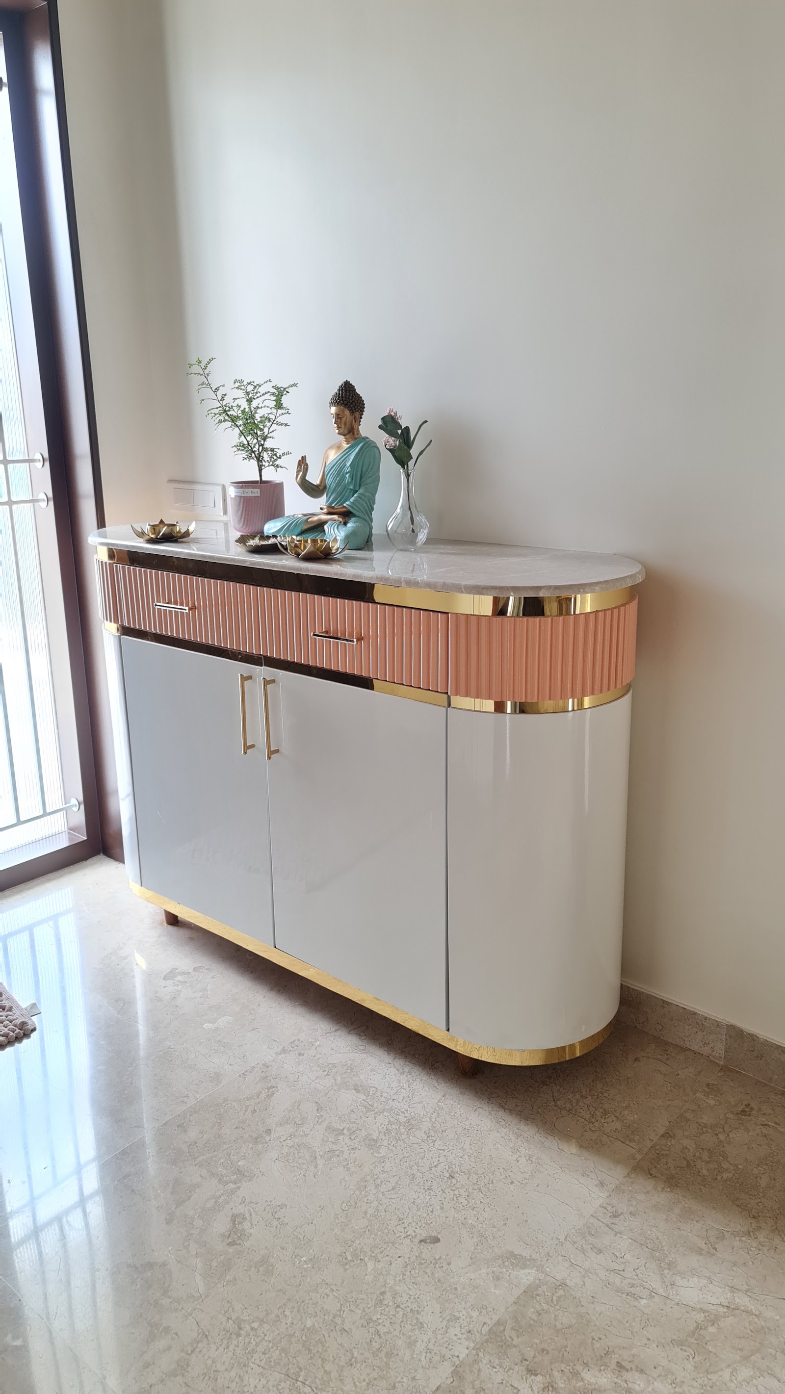 Entrance console with a curved pink-fluted and grey lacquer cabinet, brass trim, and a marble top styled with a Buddha statue and potted plant.