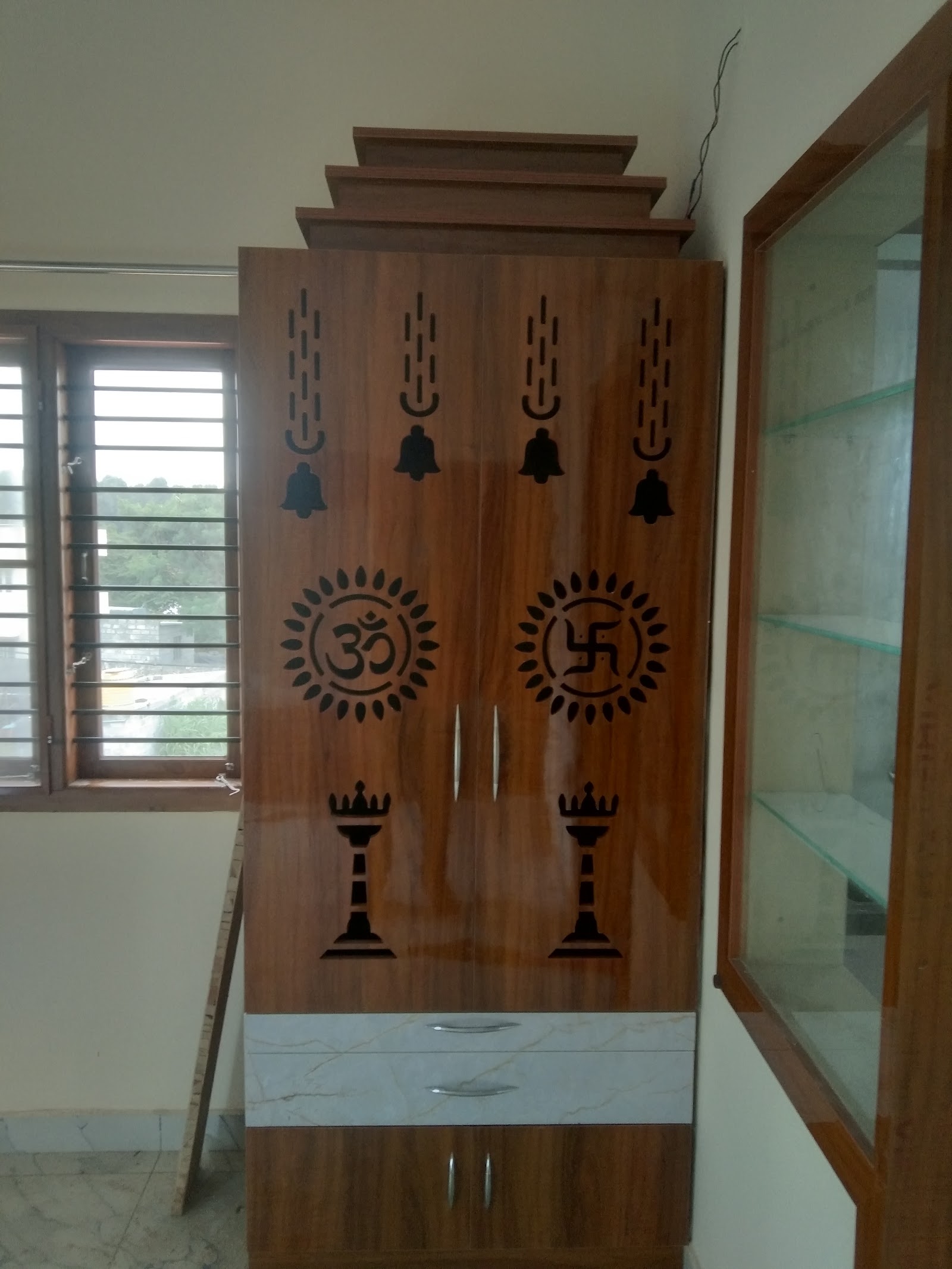 Free-standing wood-veneer pooja unit with laser-cut Om and swastik motifs, bell and diya cutouts, and a tiered pediment top, beside a marble-look drawer base.