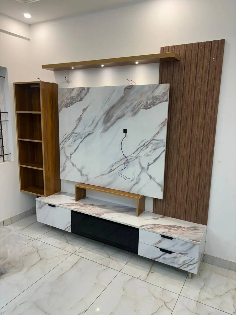 TV unit wall under installation, with a marble-look laminate feature panel and floating console flanked by a fluted wood veneer wall panel and open shelving