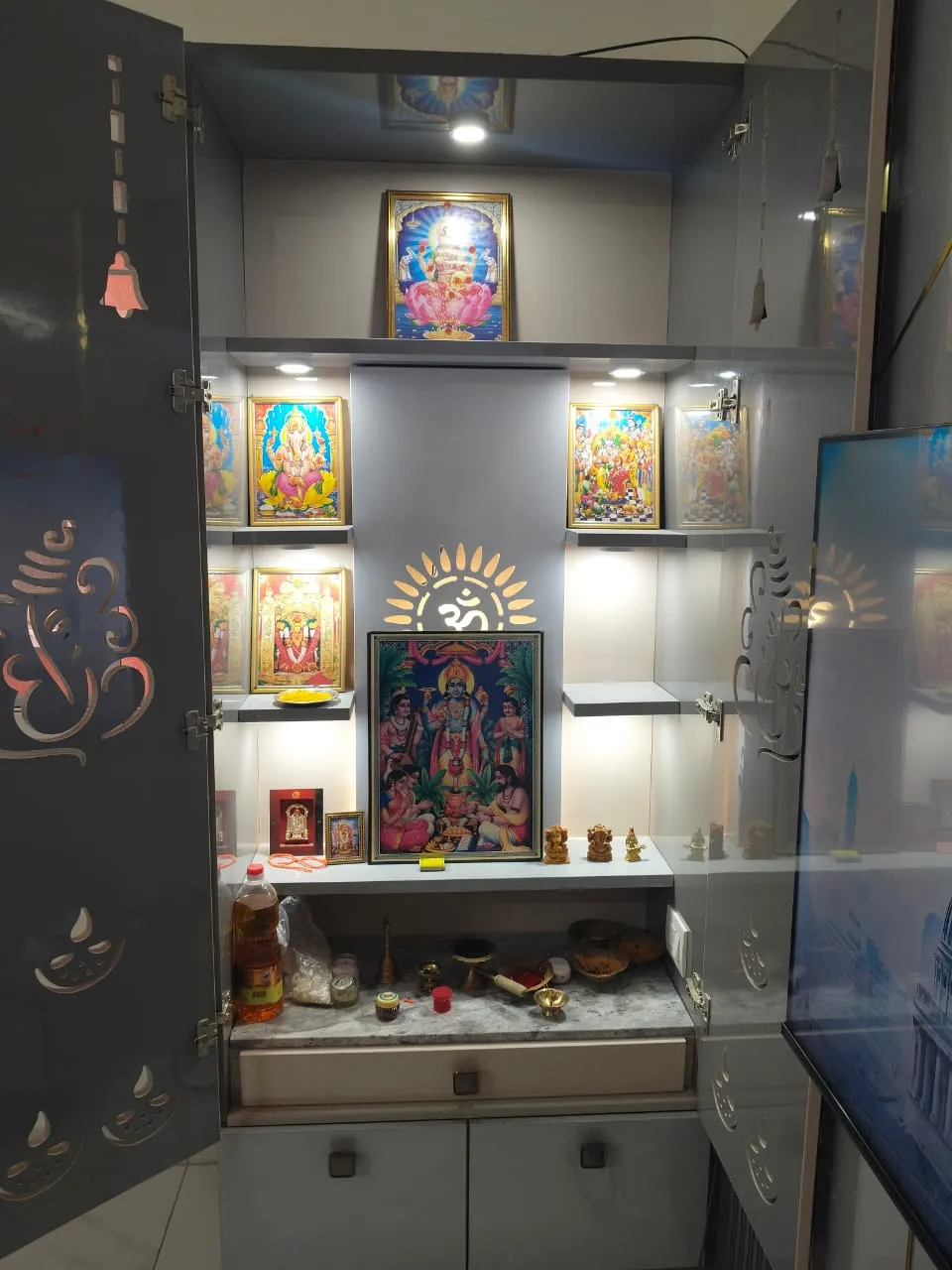 A grey lacquered pooja cabinet with laser-cut Om and diya motif doors, backlit niches displaying framed deity portraits and brass idols.